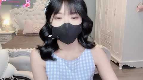 Media: Video of a young Asian woman with shoulder-length black hair and fair skin, wearing a blue and white checkered top, black mask, and headphones, in a chic, white and pink bedroom with a tufted headboard and floral decor.
