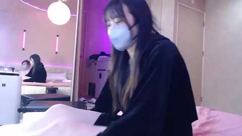 Video of a woman with long hair and a blue mask, lying in bed, with a white wall and modern decor in the background.