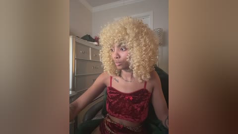 Video of a young, light-skinned woman with curly, voluminous blonde hair, wearing a red velvet crop top and matching shorts, sitting in a dimly lit room with beige furniture.