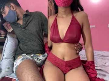 Video of a man wearing a mask and casual clothes, sitting on a bed next to a woman in a red lace lingerie set, pink mask, and gloves.