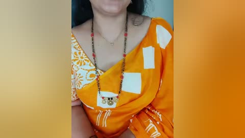 A video of a woman with light brown skin wearing a vibrant orange saree with white patterns, adorned with a black beaded necklace and white and gold pendant.
