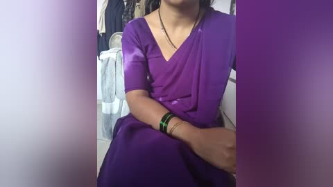 Video of a woman in a purple sari, seated in a dimly lit room with a purple gradient background. She wears a black necklace and green bracelets, and her brown hair is tied back.