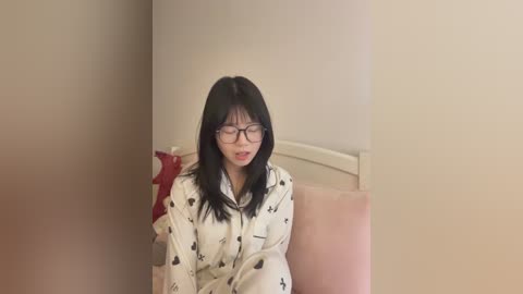 Video of an Asian woman with long black hair, wearing glasses and a white pajama set with cartoon prints, seated on a beige couch against a cream-colored wall.