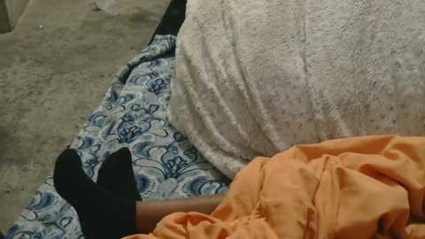 A video of a person lying on a patterned blue and white blanket, wearing black socks and orange pants, with a white, fluffy blanket beside them on a concrete floor.