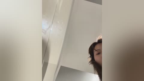 A video of a woman with long, wavy brown hair partially obscured by a white shower curtain, standing in a modern, minimalist bathroom with light-colored walls.