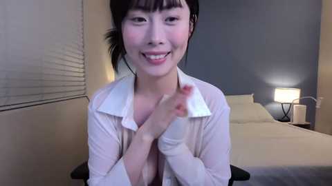 Media: Video of an Asian woman with straight black hair and bangs, wearing a white blouse, smiling and clapping hands in a modern bedroom with beige walls, a bed, and a lamp.