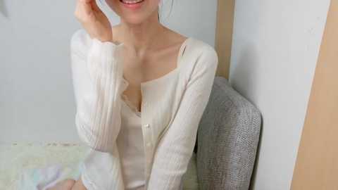 Video of a light-skinned Asian woman with a slender build, wearing a white, off-the-shoulder sweater and grey skirt, sitting on a grey chair against a white wall, smiling.