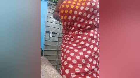 Video of a woman in a red polka-dot dress with a yellow polka-dot pattern on the left side, standing in a narrow hallway with a concrete floor and blue and pink walls.