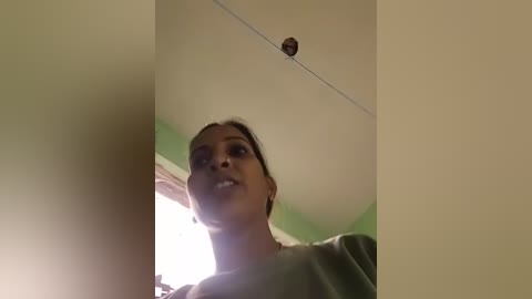 Video of a young woman with medium brown skin, dark hair tied back, wearing a green shirt, looking upwards with a slight smile, indoors with beige walls and a green ceiling, a small spider on the ceiling.
