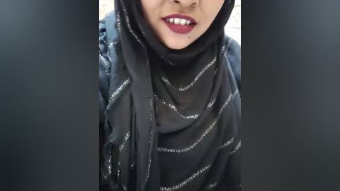 Media: Video of a woman with a dark complexion, wearing a black hijab with subtle white text, smiling with red lipstick. Background is blurred.