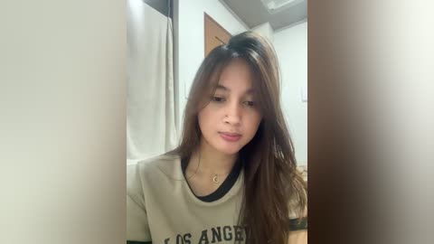 Media: Video of a young woman with long brown hair, wearing a beige t-shirt with \"Hollywood Angels\" text, indoors near a door.