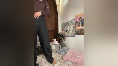 Video of a woman in a dark purple sweater and black pants, standing near a bed with pink and blue sheets. The room features posters and a white wall.
