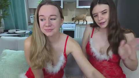 Video of two young women in red, lacy lingerie with white fur trim, smiling and gesturing in a modern kitchen with a deer statue and white cabinets.