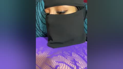 A close-up video of a person wearing a black face mask, a teal and blue patterned scarf, and a purple lace garment. The background is a gradient of blue and purple hues.
