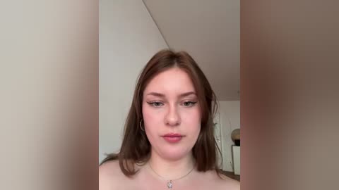 Video of a young Caucasian woman with light skin, straight brown hair, and subtle makeup, standing indoors. She wears a silver necklace. Background features a beige wall and minimal furniture.