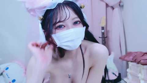 A video of an Asian woman with long black hair, wearing a pink and white bunny headband, white face mask, and a revealing black top, touching her hair in a soft, intimate indoor setting.
