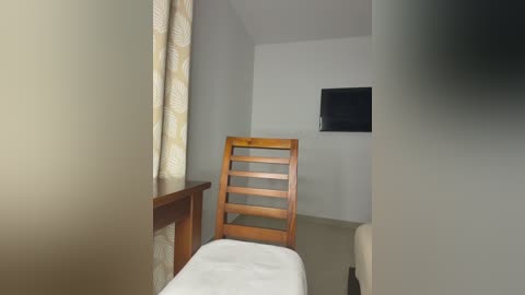 Video of a minimalist, modern room with beige walls, a wooden chair with white cushion, a small desk, and a flat-screen TV on the wall.