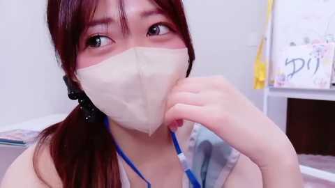 Video of an Asian woman with long brown hair in pigtails, wearing a beige surgical mask and a blue stethoscope, holding a clipboard in a clinical setting.