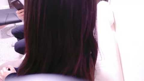 Media: Video of a person with long, straight, dark brown hair, sitting in a chair with white clothing, holding a smartphone. Background shows a light-colored carpet and part of a black leather chair.