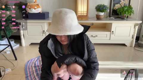 Video of a woman with a beige bucket hat, black jacket, and light skin holding a baby in a black puffer jacket, sitting on a colorful rug in a bright, modern living room with white cabinets and potted plants.