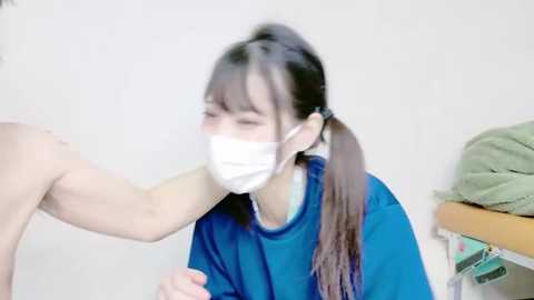 Video of an Asian woman with long dark hair in a ponytail, wearing a blue t-shirt and white surgical mask, leaning forward, touching a shirtless man's arm in a sterile medical setting.