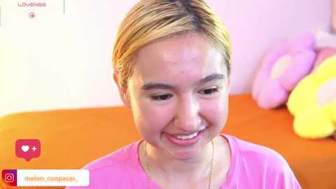 Video of a young woman with light skin, short blonde hair, and a cheerful smile, wearing a pink shirt, seated on an orange sofa with yellow and pink cushions in a brightly lit room.