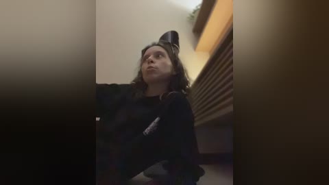 A video of a woman wearing a black cat costume, sitting on a dark carpet in a dimly lit room with beige walls and a radiator.