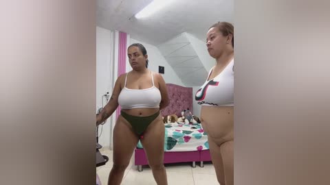 Video of two plus-sized women in lingerie, one in a white crop top and green panties, the other in a white crop top with a heart design, standing in a pink bed with stuffed animals.
