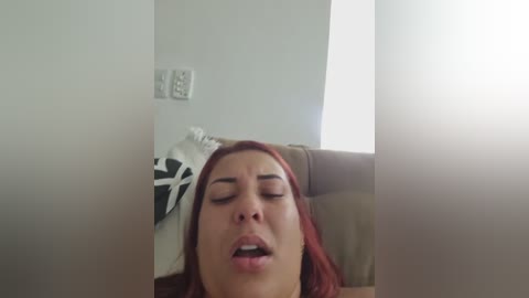 A video shows a young woman with red hair, closed eyes, and slightly open mouth, reclining on a brown leather sofa, with a white and black patterned pillow behind her.