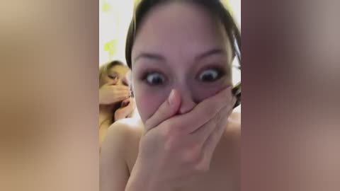 Video of a young woman with pale skin, brown hair, and wide eyes, covering her mouth with her hand, standing in a dimly lit room with blurred figures in the background.