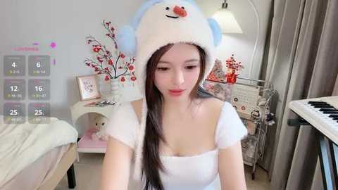 A video of a young East Asian woman with fair skin, wearing a fluffy white hat with a snowman design, and a white off-shoulder top, sitting at a piano in a cozy, decorated room.