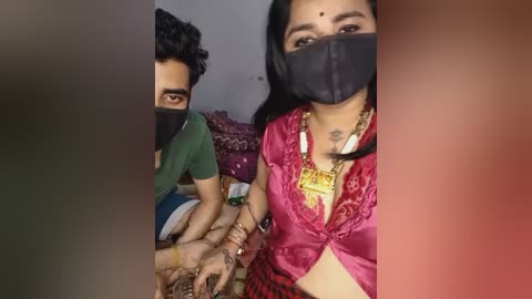 Video of two South Asian individuals, a man with curly hair and a woman with long black hair, wearing face masks, seated on a bed, possibly in a hospital setting.