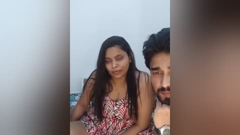 Video of a South Asian couple, woman with long black hair, red-patterned dress, resting on a bed, man with dark hair and beard, partially visible, white wall background.