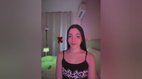 A video of a young woman with long black hair, wearing a black tank top with white floral designs, standing in a dimly lit room with soft lighting and a Christmas tree in the background.