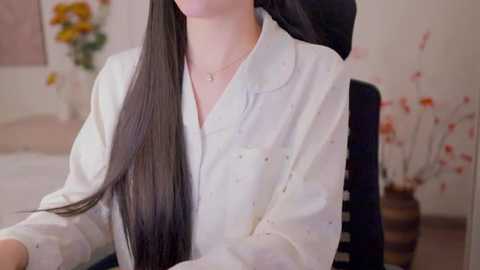 A video of a woman with long, straight black hair, wearing a white button-up shirt, seated in a black office chair. The background features a blurred, modern office setting with white walls and floral decorations.