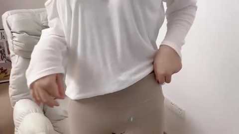 A video of a person lifting a white long-sleeved shirt, revealing a belly button piercing and a tattoo on their lower abdomen, standing in a room with a white chair and a shelf in the background.