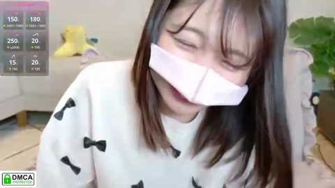 A video shows an Asian woman with shoulder-length black hair, wearing a white sweatshirt with black bows, gagged with white tape, sitting on a floor in a living room.