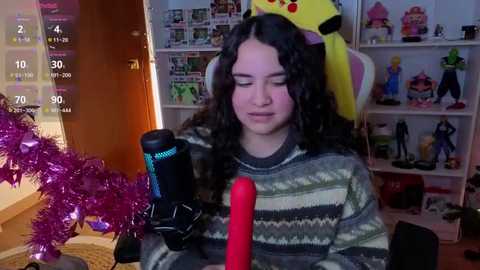 Video of a young woman with curly dark hair, wearing a striped sweater, holding a pink vibrator, in a cluttered bedroom with toys and a pink tinsel garland.