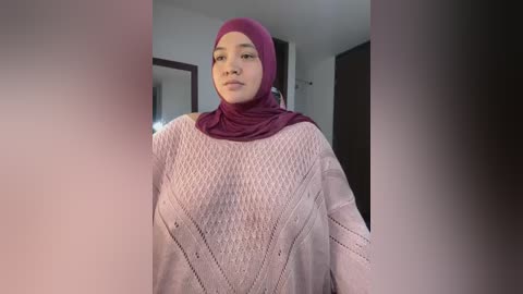 A video of a light-skinned woman with a round face, wearing a purple hijab and a light pink, knitted sweater with diamond patterns, standing indoors in a modern, minimalist setting.