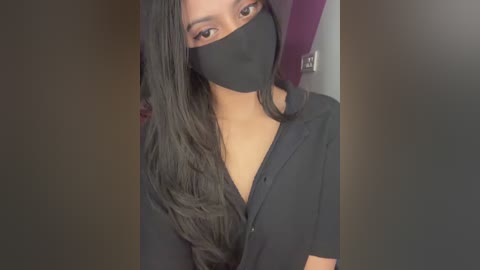 A video of a young woman with long, dark hair, wearing a black mask and a black shirt, standing against a dark background.