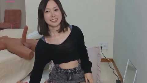Media: A video of a young Asian woman with shoulder-length black hair, wearing a black crop top and denim shorts, smiling, sitting on a beige carpet in a minimalist living room with a white wall and electrical outlets.
