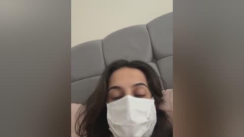 A video of a person with long dark hair, wearing a white surgical mask, lying on a gray upholstered headboard, with blurred background.