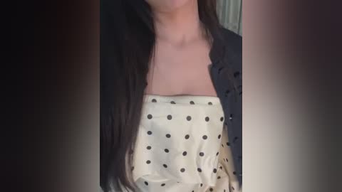 Video of a woman with long, dark hair, wearing a black jacket over a white blouse with black polka dots. The image is dimly lit, with blurred background.