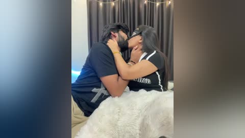 Video of a couple kissing passionately on a bed with white fur blanket. He has a beard and wears a black shirt, she has long black hair, wears a black shirt, and touches his face. Dark curtains and string lights in the background.