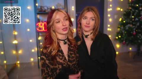 Video of two women with long hair, wearing leopard print dresses and black chokers, standing indoors near a decorated Christmas tree with fairy lights.