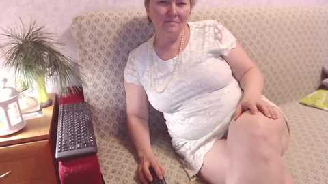 Video of a middle-aged, light-skinned woman with short brown hair, wearing a white lace dress, sitting on a beige-patterned couch, holding a laptop on her lap, in a cozy living room.