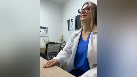 A video of a young, blonde, light-skinned woman in glasses, wearing a white lab coat over a blue shirt, sitting at a desk in an optometrist's office.