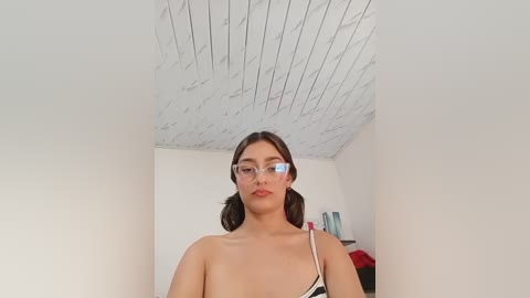 Video of a young woman with light skin, brown hair, and glasses, wearing a black and white dress, standing in a minimalist room with white walls and a wooden ceiling.