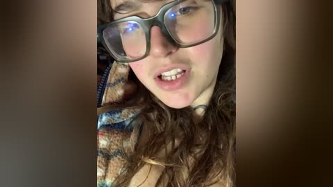 A close-up video of a young woman with long, wavy brown hair wearing large, black, rectangular glasses, a plaid jacket, and a slightly open mouth, showing her teeth. The background is blurred and dark.