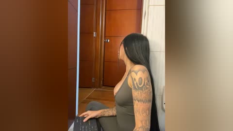 Video of a tattooed woman with long black hair, wearing a sleeveless gray top, sitting on the floor near a wooden door in a tiled hallway.